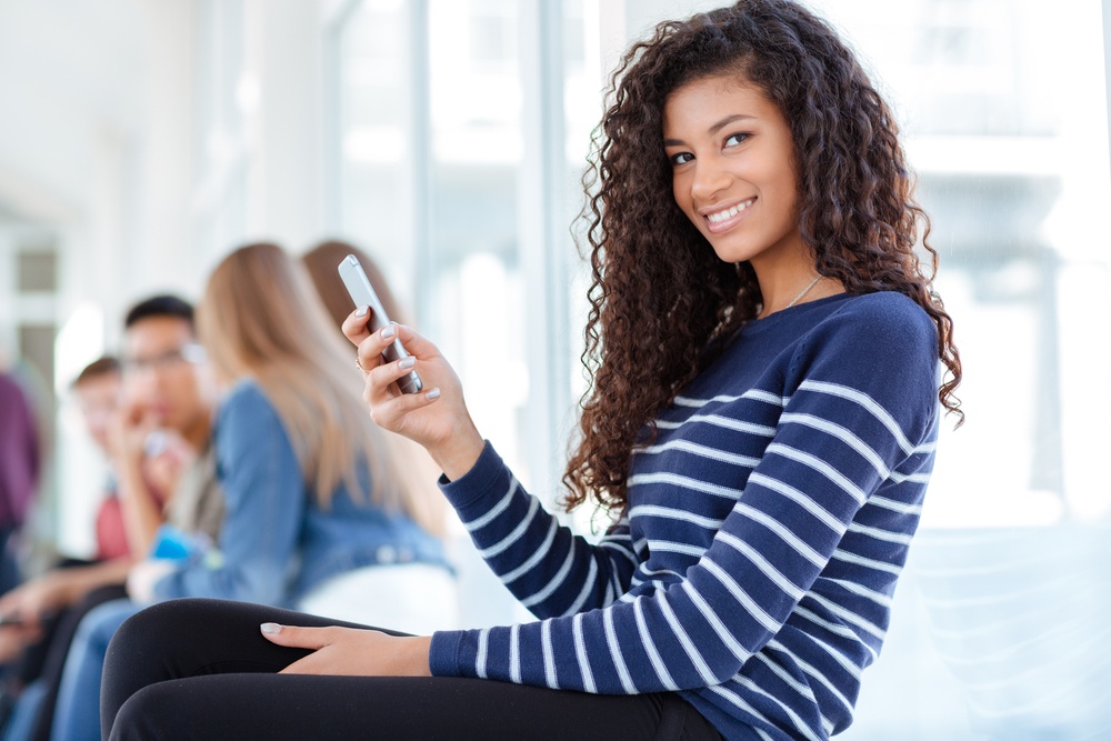 Portrait Of A Happy Afro American Female Student Using Smartphone In University Hall And Looking At Camera